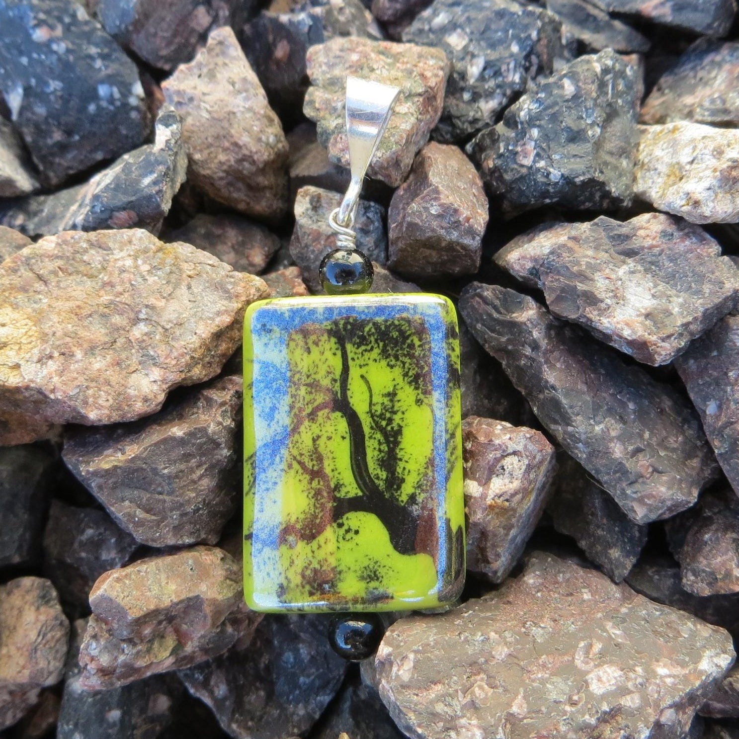 May include: A rectangular pendant with a silver bail and a black bead. The pendant features a lime green background with a black tree design and blue accents. The pendant is displayed on a bed of rocks.