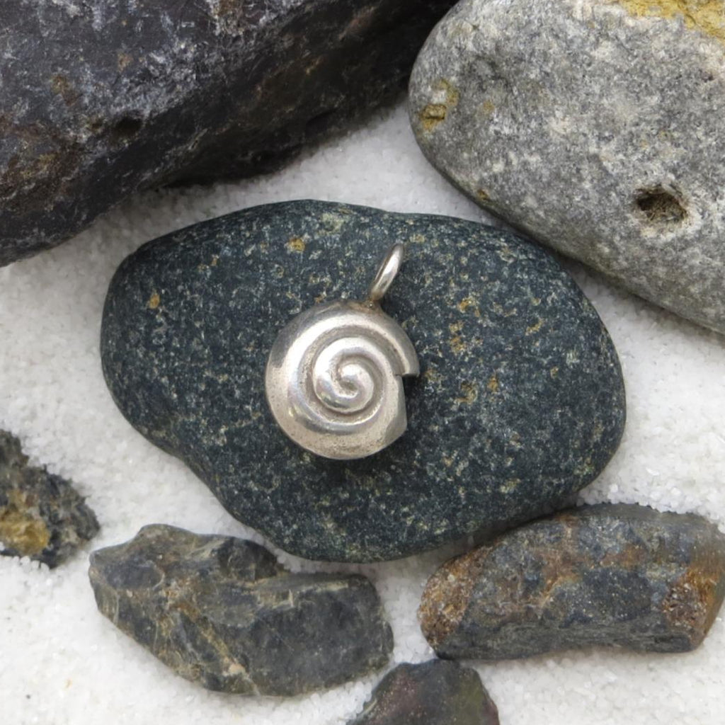 May include: A silver pendant with a spiral design, resting on a dark gray stone. The pendant is surrounded by various rocks and a white, sandy surface. The spiral design is a prominent feature of the pendant.