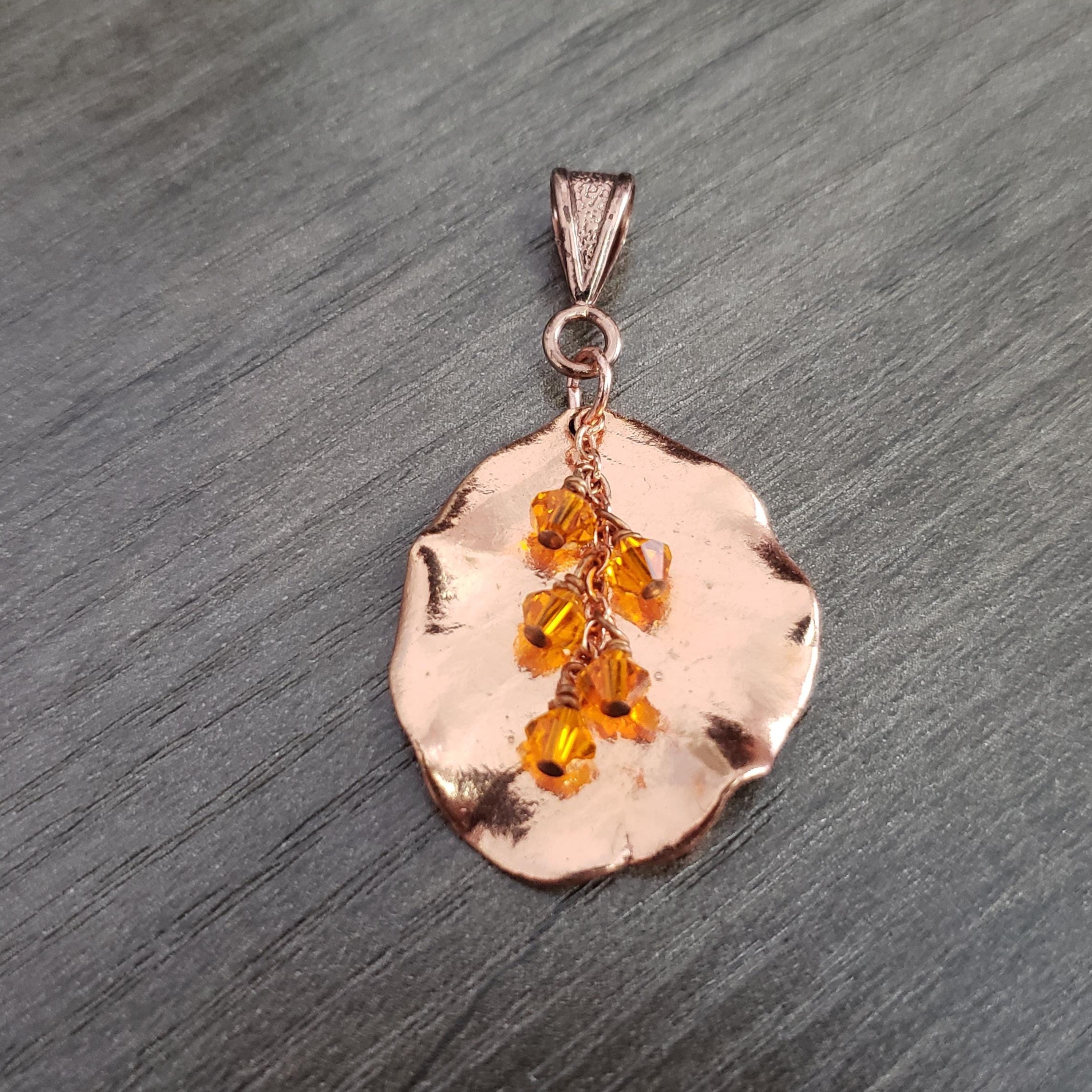 May include: A rose gold pendant with a leaf-like shape. Dangling from the pendant are several orange crystal beads. The pendant has a textured surface and a small loop at the top for a chain.