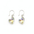 May include: A pair of heart-shaped earrings with iridescent crystals. The earrings are suspended from silver-colored hooks. The crystals reflect yellow, blue, and green hues. The earrings are displayed on a textured, gray stone surface.