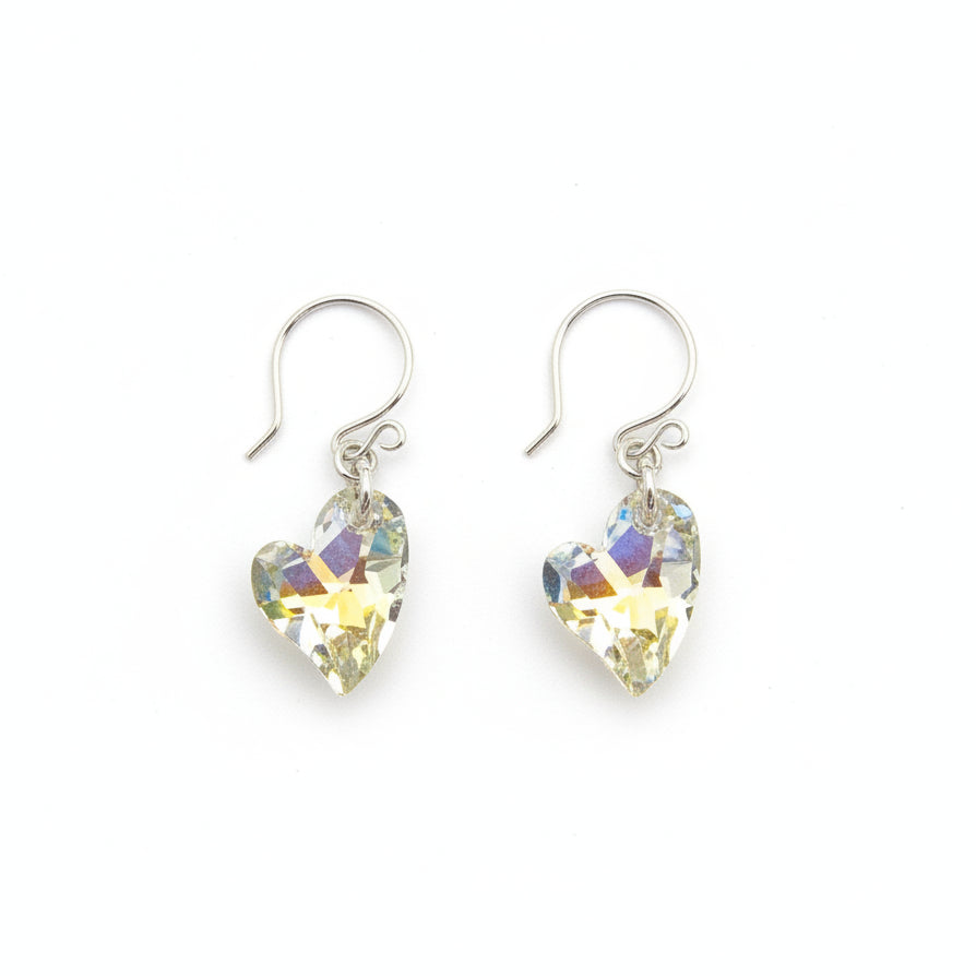 May include: A pair of heart-shaped earrings with iridescent crystals. The earrings are suspended from silver-colored hooks. The crystals reflect yellow, blue, and green hues. The earrings are displayed on a textured, gray stone surface.