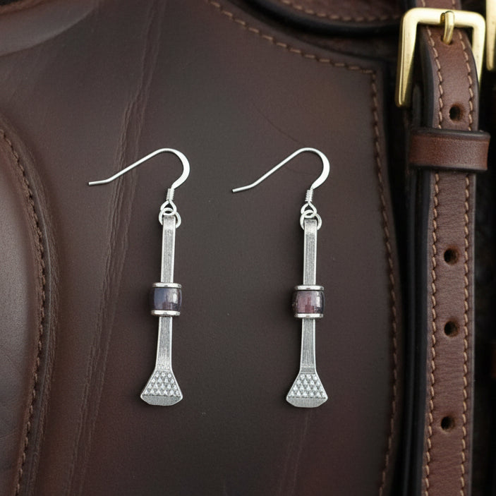 May include: A pair of silver-toned earrings with a unique design. Each earring features a long, slender bar with a small, rectangular purple bead in the center. The bottom of each earring has a textured, flared shape. The earrings hang from a simple hook.