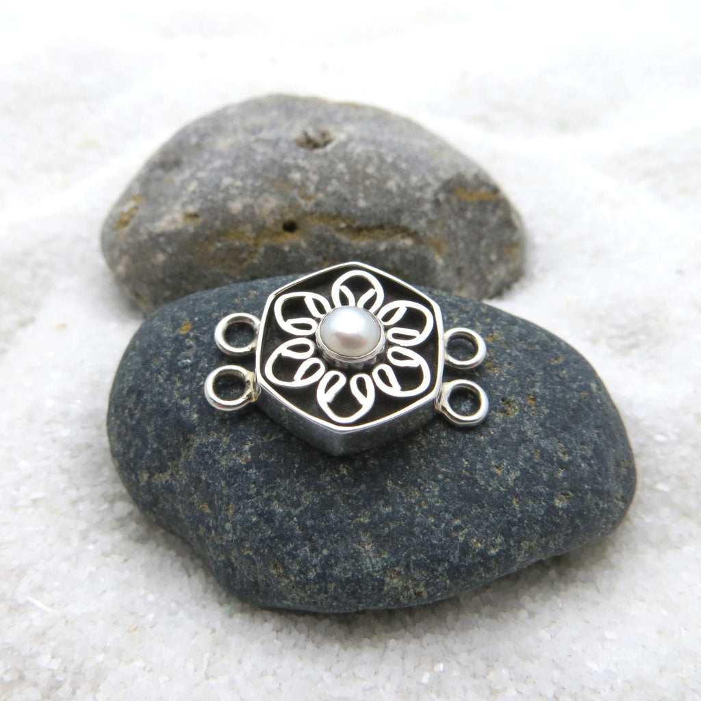 May include: A silver-colored jewelry clasp with a floral design and a central pearl. The clasp is hexagonal with a flower-shaped cutout pattern. It has two loops on each side for attachment. The clasp is resting on a dark gray stone.