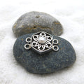 May include: A silver-colored jewelry clasp with a floral design and a central pearl. The clasp is hexagonal with a flower-shaped cutout pattern. It has two loops on each side for attachment. The clasp is resting on a dark gray stone.