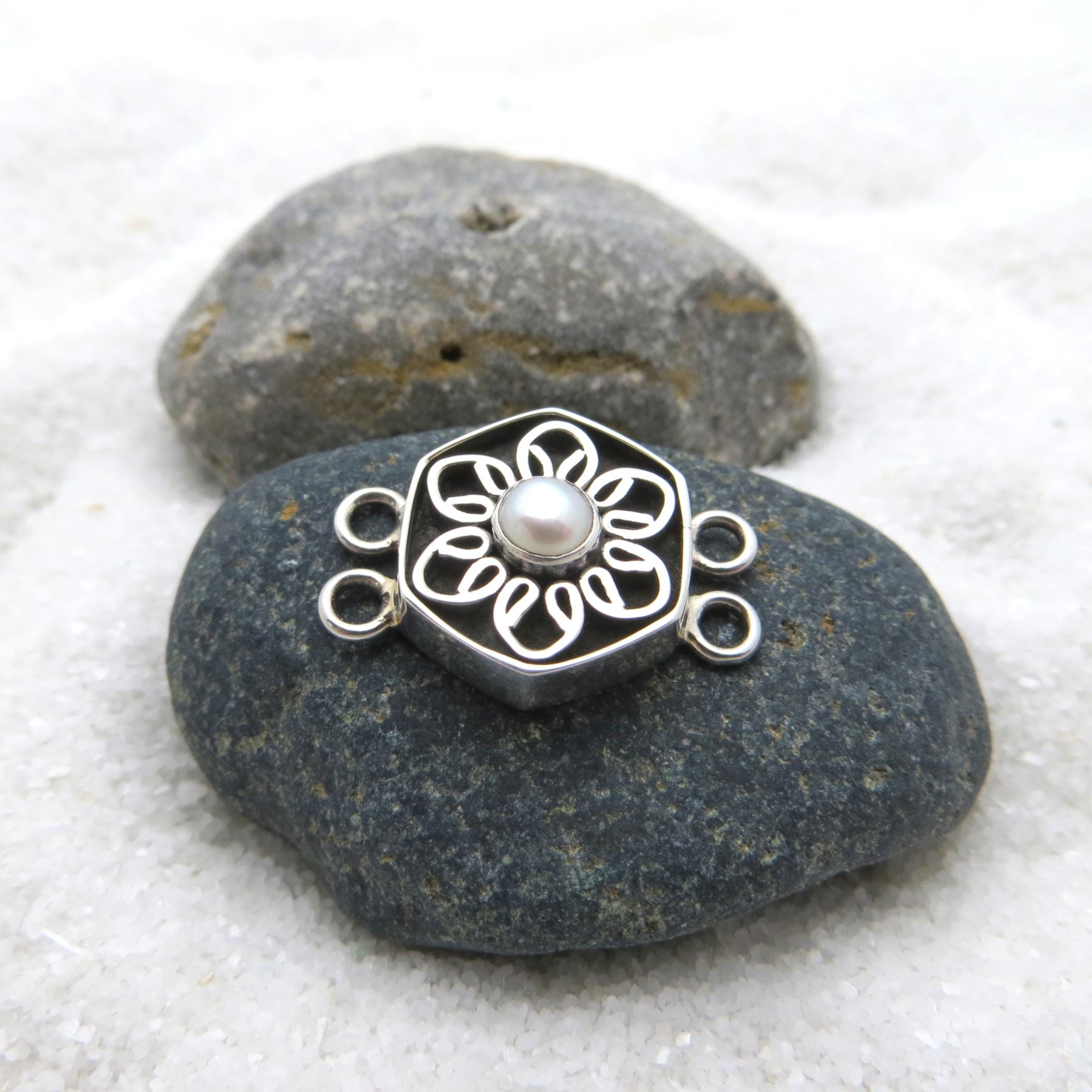 May include: A silver-colored jewelry clasp with a floral design and a central pearl. The clasp is hexagonal with a flower-shaped cutout pattern. It has two loops on each side for attachment. The clasp is resting on a dark gray stone.