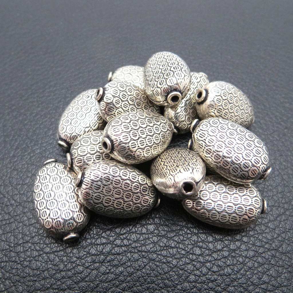 May include: A collection of silver-colored, oval-shaped beads. Each bead has a detailed, repeating pattern and a hole at each end. The beads are scattered on a dark, textured surface, creating a contrast that highlights their intricate design.