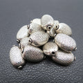 May include: A collection of silver-colored, oval-shaped beads. Each bead has a detailed, repeating pattern and a hole at each end. The beads are scattered on a dark, textured surface, creating a contrast that highlights their intricate design.