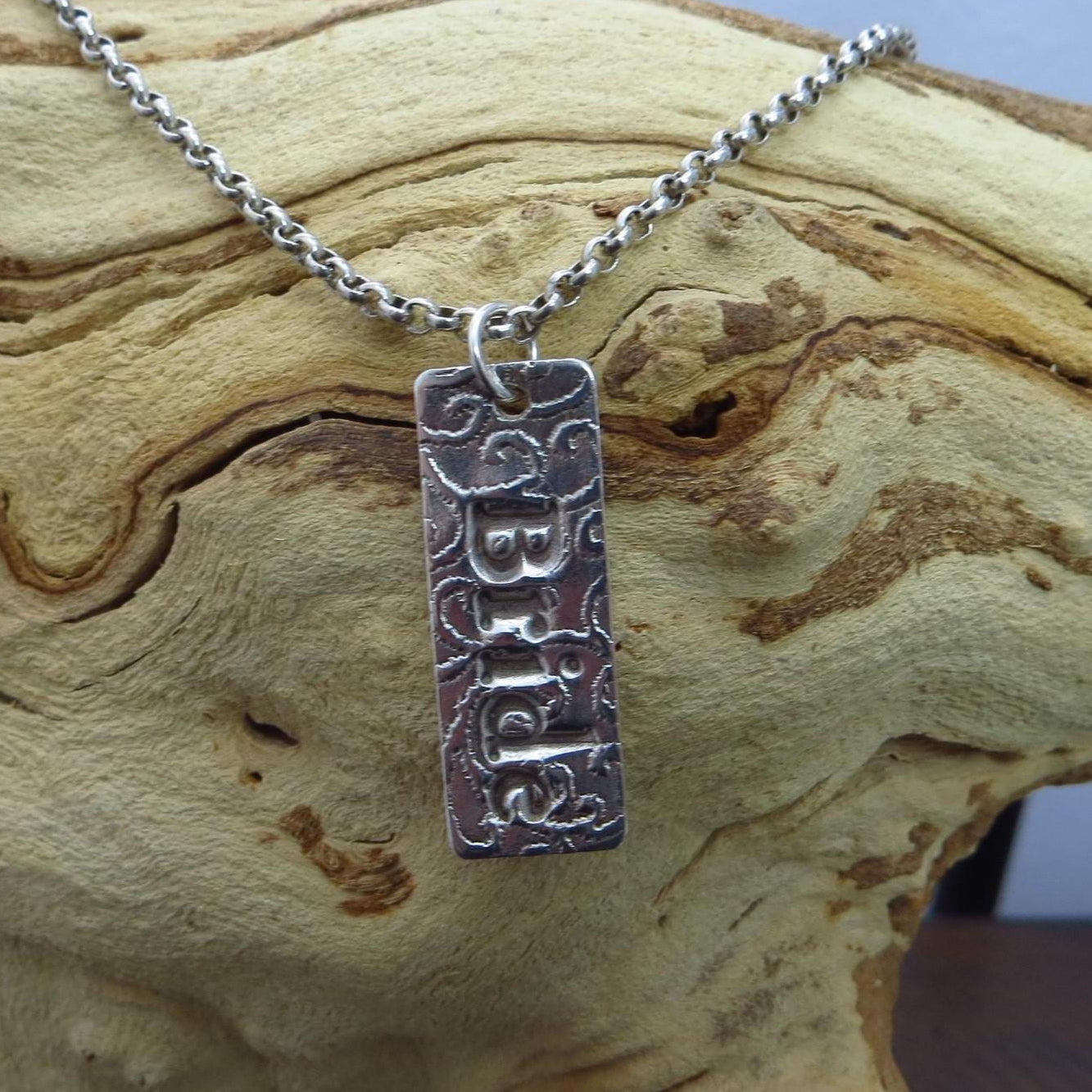 May include: A silver-toned pendant necklace with the name "Bria" engraved on a rectangular charm. The charm has a textured background and is suspended from a delicate chain. The necklace is displayed against a natural wood background.