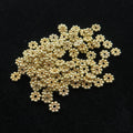 May include: A collection of small, gold-colored metal flower-shaped beads. Each bead has a central hole surrounded by a ring of smaller, spherical beads, creating a floral design. The beads are scattered on a black surface.