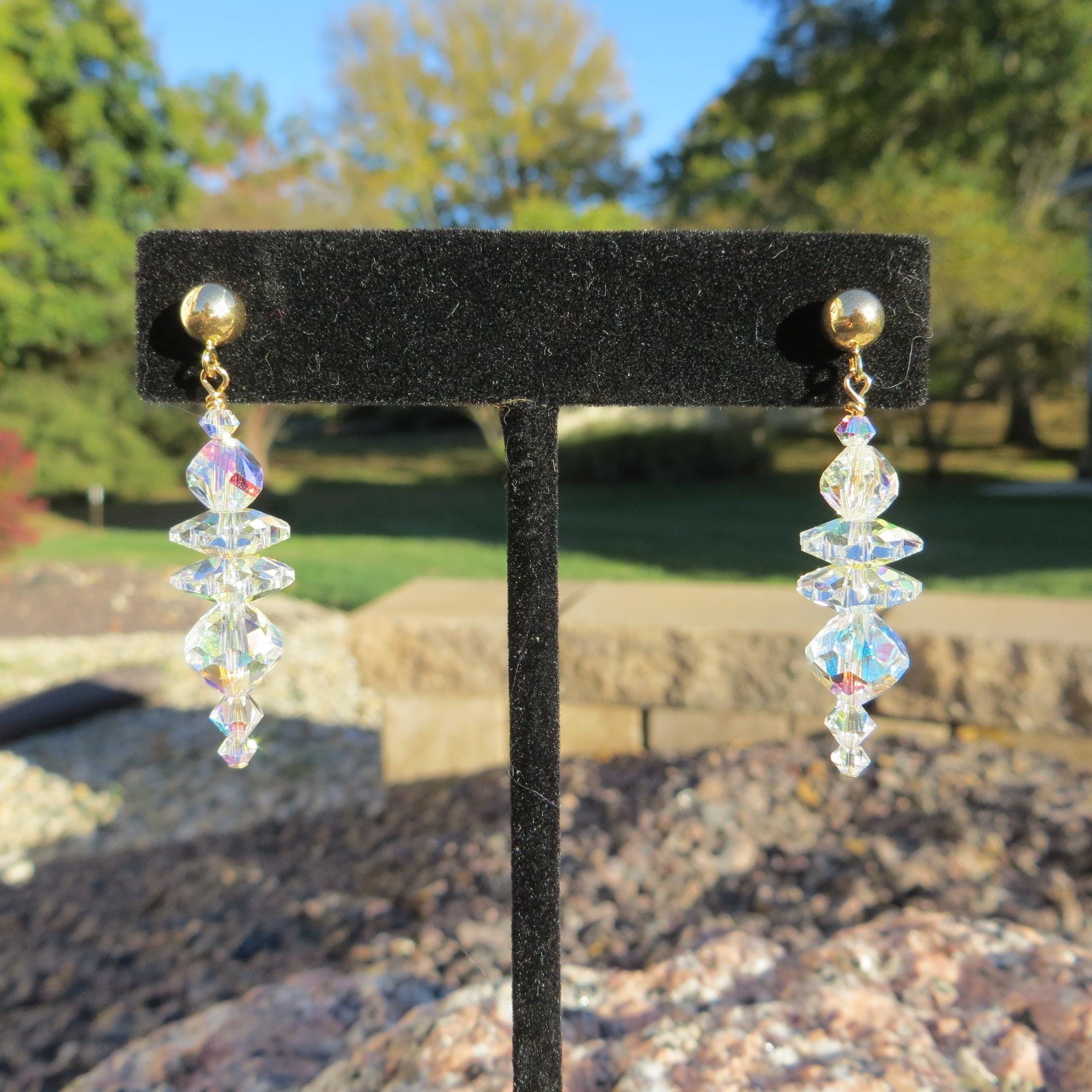 May include: A pair of dangle earrings with gold-colored studs and clear, iridescent crystal beads. The crystals are arranged in a cascading design, reflecting light. The earrings are displayed on a black velvet stand.