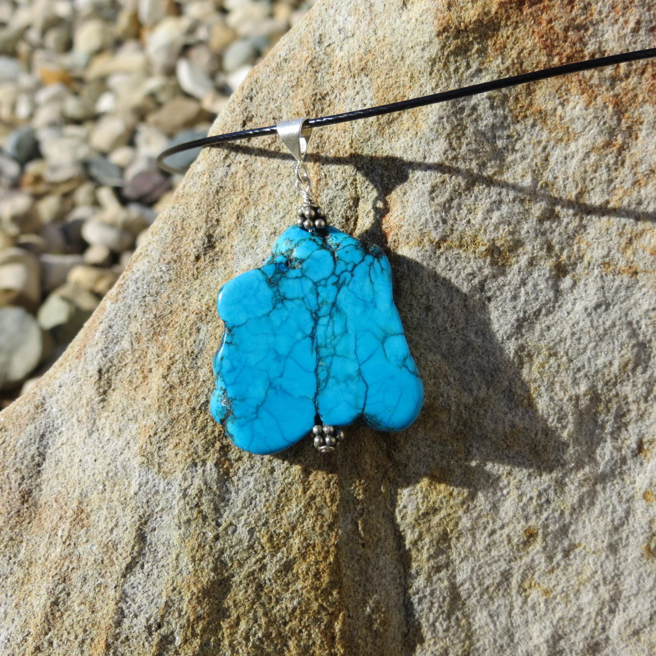 May include: A turquoise pendant necklace with a silver bail and black cord. The pendant is a large, irregularly shaped stone with a vibrant blue color and intricate veining. The necklace is displayed on a textured, light brown stone surface.